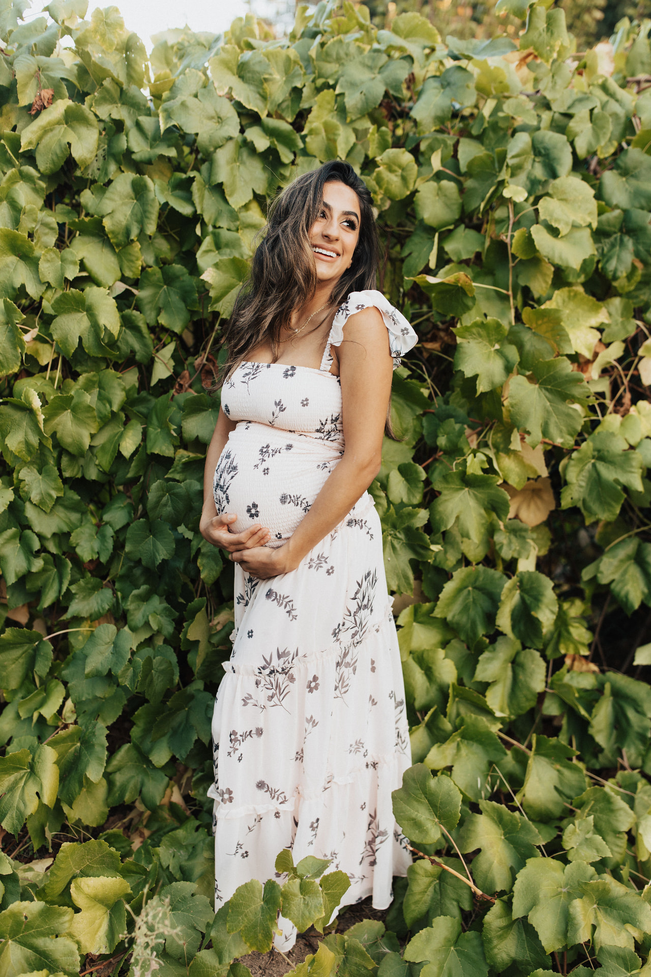 Smiling pregnant woman in a cream floral dress cradling her bump against a wall of green leaves